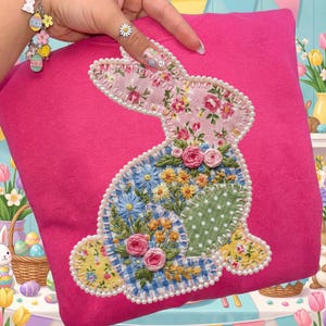 May include: A bright pink sweatshirt featuring a patchwork bunny design. The bunny is adorned with floral patterns, including pink roses, blue and yellow flowers, and a green polka dot patch. The design is outlined with white pearls.