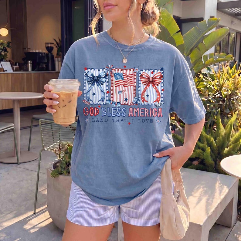 Puede incluir: Camiseta azul con un dise&ntilde;o patri&oacute;tico con lazos y una bandera estadounidense. El texto "GOD BLESS AMERICA" y "LAND THAT I LOVE" est&aacute; impreso en la camiseta. La persona lleva pantalones cortos a rayas azules y blancas y sostiene una bebida de caf&eacute;.