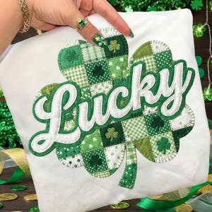 May include: A white fabric item featuring a large shamrock design in green and white patchwork. The word "Lucky" is embroidered in white script across the shamrock. The shamrock design incorporates various green patterns, including polka dots and gingham.