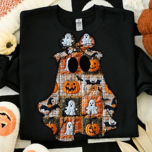 May include: Black sweatshirt with a patchwork ghost design. The ghost is made of orange, white, and black fabric squares, featuring pumpkins, bats, and ghost figures. The ghost has two large black eye holes and a bow.