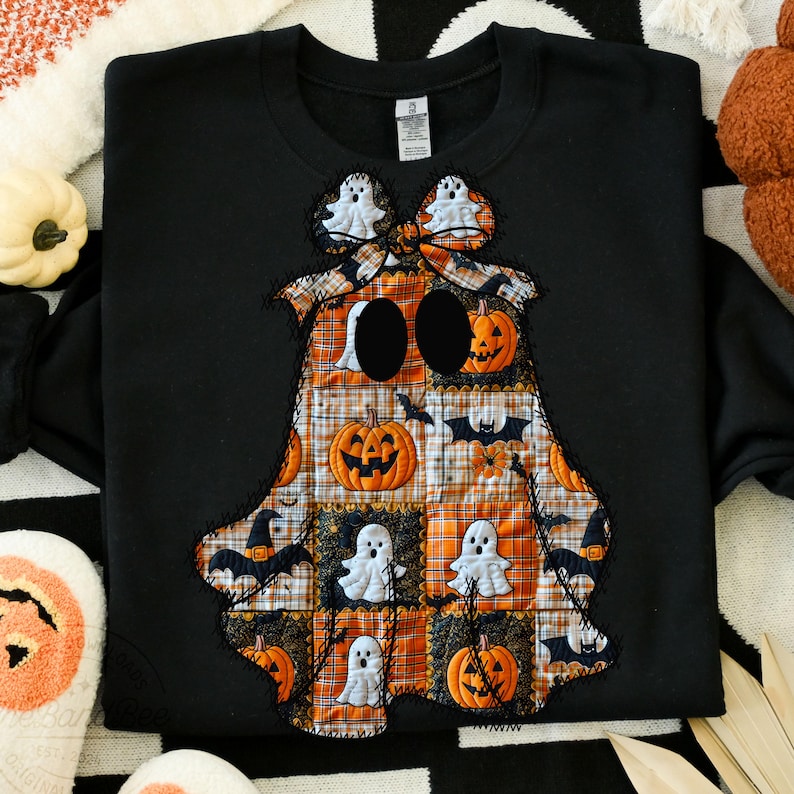 May include: Black sweatshirt with a patchwork ghost design. The ghost is made of orange, white, and black fabric squares, featuring pumpkins, bats, and ghost figures. The ghost has two large black eye holes and a bow.