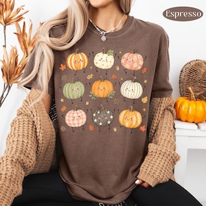 Puede incluir: Una camiseta color marr&oacute;n espresso con un estampado gr&aacute;fico de nueve calabazas de dibujos animados con caras sonrientes, brazos y piernas. Las calabazas tienen varios patrones como rayas, puntos y estrellas, intercaladas con peque&ntilde;as hojas y bellotas.