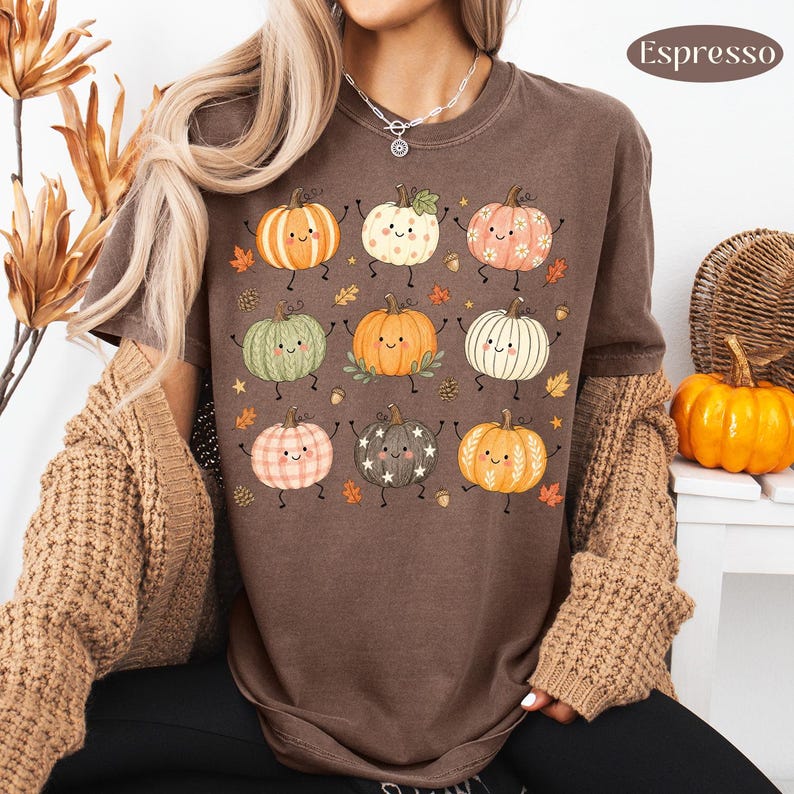 Puede incluir: Una camiseta color marr&oacute;n espresso con un estampado gr&aacute;fico de nueve calabazas de dibujos animados con caras sonrientes, brazos y piernas. Las calabazas tienen varios patrones como rayas, puntos y estrellas, intercaladas con peque&ntilde;as hojas y bellotas.