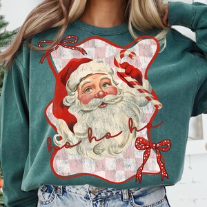 May include: Teal green sweatshirt featuring a vintage Santa Claus graphic. The Santa design includes a red hat, white beard, and a candy cane. The words "ho ho ho" are written in red script. Red bows with white polka dots accent the design.