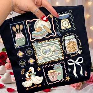 May include: Black t-shirt featuring a charming, illustrated design of kitchen-themed elements. The design includes a teapot, teacups, jars of cookies and jam, a scale, and ducks. The artwork is framed with decorative borders and floral accents.