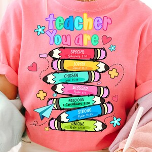 May include: Pink t-shirt with the words "teacher You are" in colorful letters. Below the text are stacked pencils with words like "Special," "Lovely," and "Chosen." Each pencil has a Bible verse reference. The design includes hearts, flowers, and a paper airplane.