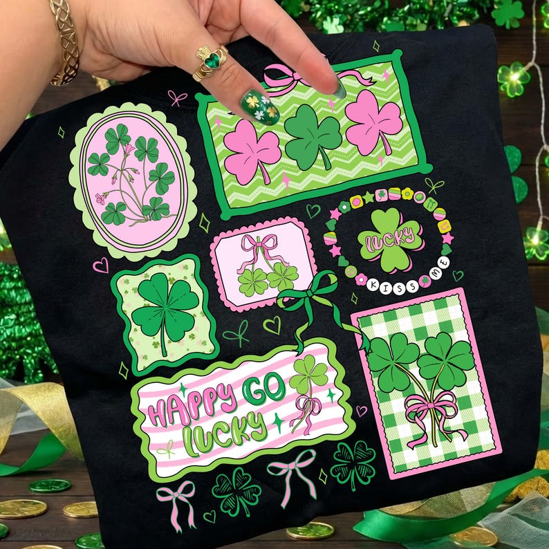 Op de afbeelding: Zwart T-shirt met St. Patrick's Day-thema graphics. Het ontwerp bevat klavers, strikken en de zinnen "Happy Go Lucky" en "Kiss Me". De graphics zijn in groen, roze en wit.