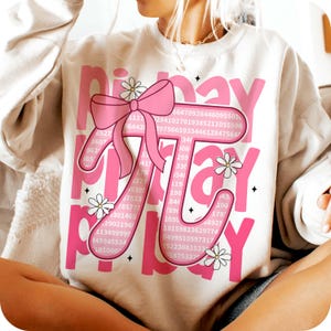 May include: A white sweatshirt with a pink graphic design that says "Pi Day" with a pink bow and a pi symbol made up of numbers.