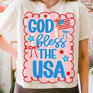 Puede incluir: Camiseta color crema con un dise&ntilde;o patri&oacute;tico. El gr&aacute;fico incluye el texto "GOD bless THE USA" en azul, con un fondo de rayas rojas y blancas, estrellas y una peque&ntilde;a bandera estadounidense.