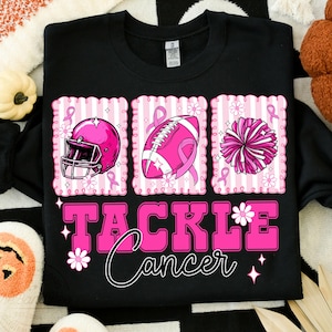 May include: Black sweatshirt with a pink graphic that says "Tackle Cancer." The graphic includes a pink football helmet, football, and pom-pom, each with a pink ribbon. White flowers and stars accent the design.