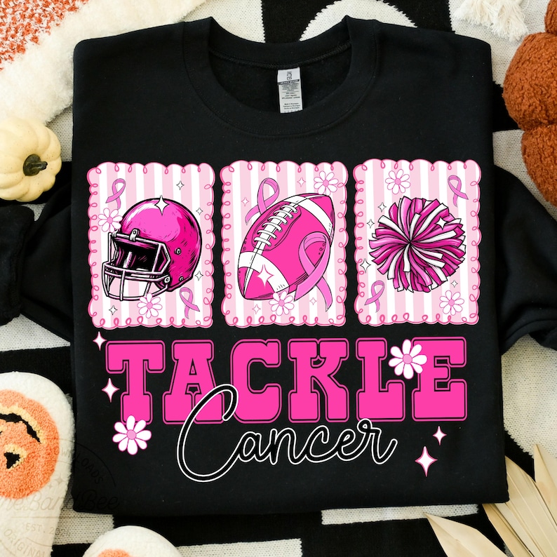 May include: Black sweatshirt with a pink graphic that says "Tackle Cancer." The graphic includes a pink football helmet, football, and pom-pom, each with a pink ribbon. White flowers and stars accent the design.