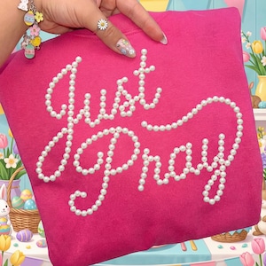 Op de afbeelding: Een felroze sweatshirt met de woorden "Just Pray" uitgespeld in witte parel kralen. De sweatshirt is effen van kleur en heeft een eenvoudig, klassiek ontwerp. De afbeelding is goed belicht en de tekst is gemakkelijk te lezen.