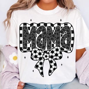 May include: White t-shirt with a black and white chequered design featuring the word "Mama" in a black and white leopard print. A black and white football bow is below the word.