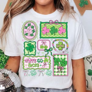 Op de afbeelding: Wit T-shirt met een St. Patrick's Day ontwerp. Het ontwerp bevat omlijste klavers in groen en roze, en de woorden "Happy Go Lucky". Een casual kledingstuk, perfect om St. Patrick's Day te vieren.