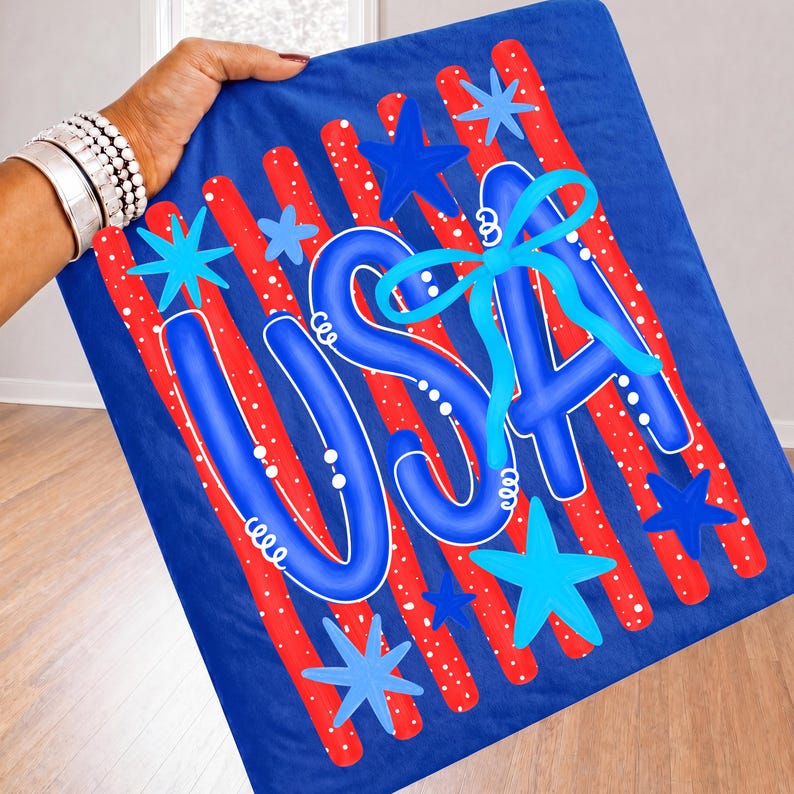 May include: A blue blanket with the word "USA" in blue script, accented with a light blue bow. Red stripes with white dots and blue stars decorate the blanket, creating a patriotic theme.