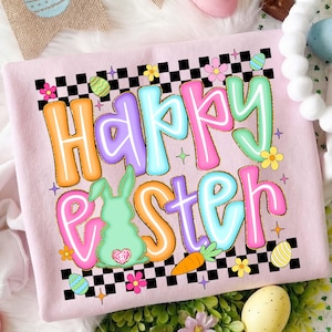 May include: A pale pink sweatshirt with a vibrant "Happy Easter" design. The graphic includes a bunny, carrot, and Easter eggs, framed by a black and white checkered pattern. The text is in shades of orange, pink, and blue.