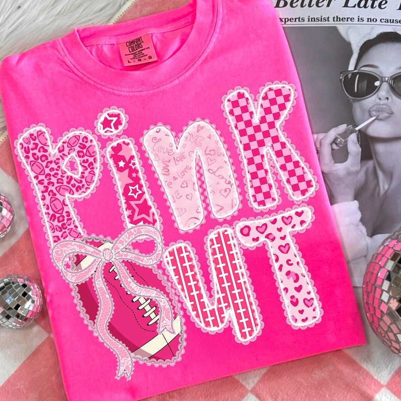 May include: Bright pink t-shirt with the words "PINK OUT" in a decorative font. The letters are embellished with various patterns, including leopard print, hearts, and a checkered design. A pink football with a bow is featured below the word "PINK".