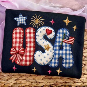 K&ouml;nnte beinhalten: Ein marineblaues Textil mit dem Wort "USA" in rot-wei&szlig; karierten, wei&szlig; gepunkteten und blau-wei&szlig; karierten Buchstaben. Das Design umfasst Sterne, eine Schleife, ein Herz und eine kleine amerikanische Flagge. Das Gesamtthema ist patriotisch.