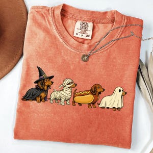 Peut inclure: Un t-shirt couleur corail orn&eacute; d'un motif repr&eacute;sentant quatre teckels d&eacute;guis&eacute;s pour Halloween : une sorci&egrave;re, une momie, un hot-dog et un fant&ocirc;me. Le t-shirt pr&eacute;sente un col rond, une &eacute;tiquette Comfort Colors et une fine cha&icirc;ne argent&eacute;e.