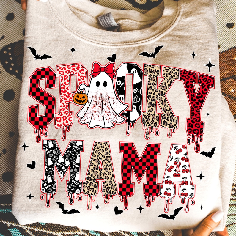 May include: A beige sweatshirt with the words "Spooky Mama" in a mix of fonts and patterns. The word "Spooky" has a checkered pattern, leopard print, and dripping blood effect. A ghost with a bow and pumpkin is in the center. Bats and stars are scattered around.