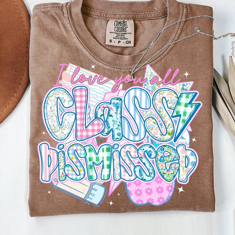 May include: A brown t-shirt with the text "I love you all Class Dismissed" in a colorful patchwork style. The text is surrounded by stars, a lightning bolt, and a pencil.