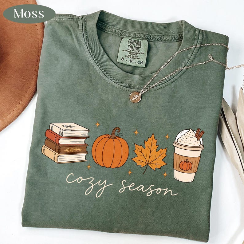 May include: A moss green t-shirt featuring a graphic print of a stack of books, an orange pumpkin, a maple leaf, and a coffee cup with whipped cream. The text "cozy season" is printed below the graphics in a white cursive script font.