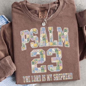 May include: A brown sweatshirt with the text "PSALM 23" in a patchwork floral design. The phrase "THE LORD IS MY SHEPHERD" is printed at the bottom. A silver necklace with a round pendant hangs on the neckline.