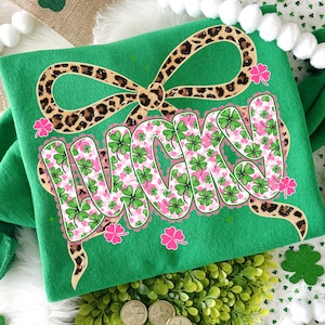 May include: Green t-shirt with the word "LUCKY" in a bubble letter style, filled with green shamrocks and pink floral accents. A leopard print bow is above the word, and pink shamrock accents are scattered around the design.