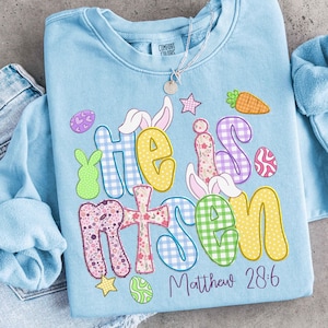May include: Light blue sweatshirt with the words "He is Risen" in colorful, patterned letters. The design includes Easter-themed graphics like bunnies, eggs, and a carrot. The text "Matthew 28:6" is printed below the main text. A silver necklace is visible.