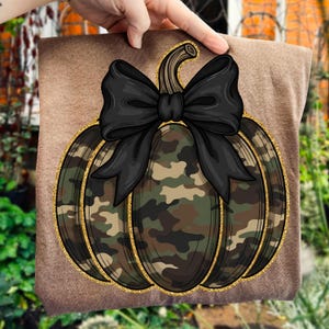 May include: A brown t-shirt featuring a pumpkin design. The pumpkin has a camouflage pattern in shades of green, brown, and tan, outlined with gold glitter. A large black bow sits atop the pumpkin, adding a decorative touch. The design is centered on the shirt.