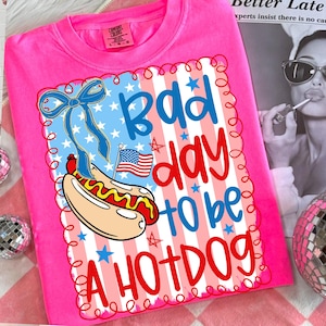 May include: Bright pink t-shirt with a graphic that says "Bad day to be a Hotdog." The design includes a hotdog, American flag, and a blue bow. The shirt is a Comfort Colors brand.