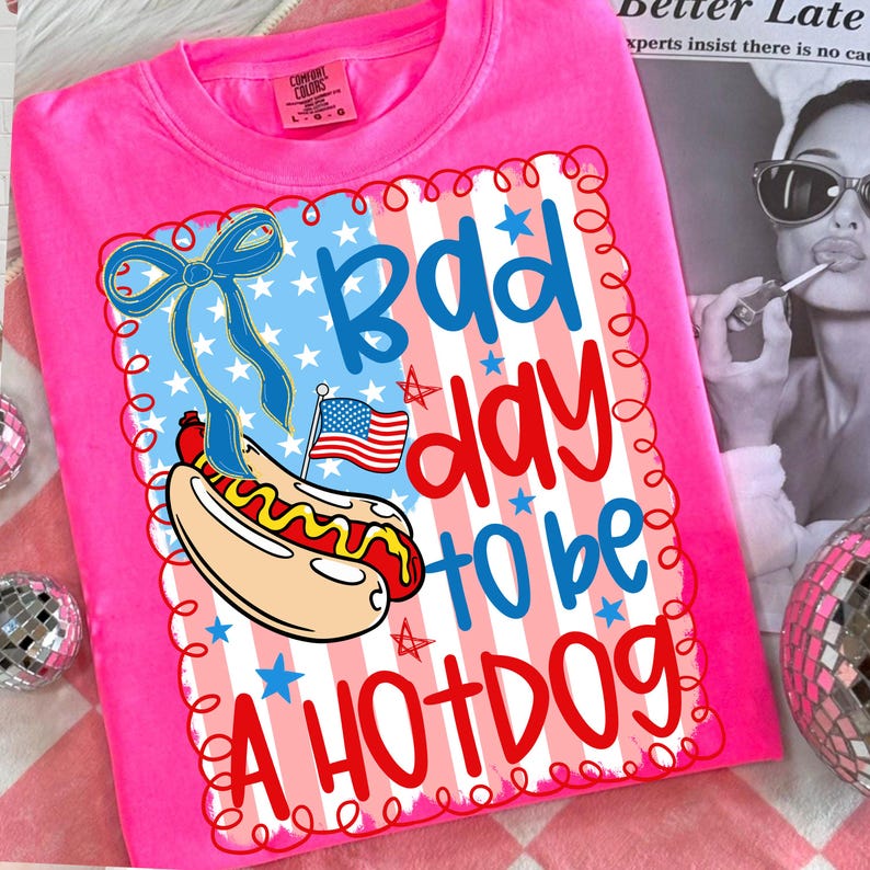 May include: Bright pink t-shirt with a graphic that says "Bad day to be a Hotdog." The design includes a hotdog, American flag, and a blue bow. The shirt is a Comfort Colors brand.