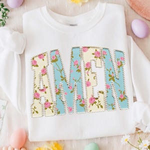 May include: White sweatshirt with the word "AMEN" in large, decorative letters. The letters are outlined with small, silver beads and feature a floral pattern in pink, yellow, and blue. The sweatshirt is a casual, comfortable style.