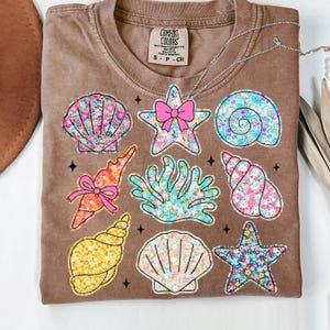 May include: A brown t-shirt with a floral pattern design featuring seashells, starfish, and coral. The text "COMFORT COLORS" is printed on the shirt.