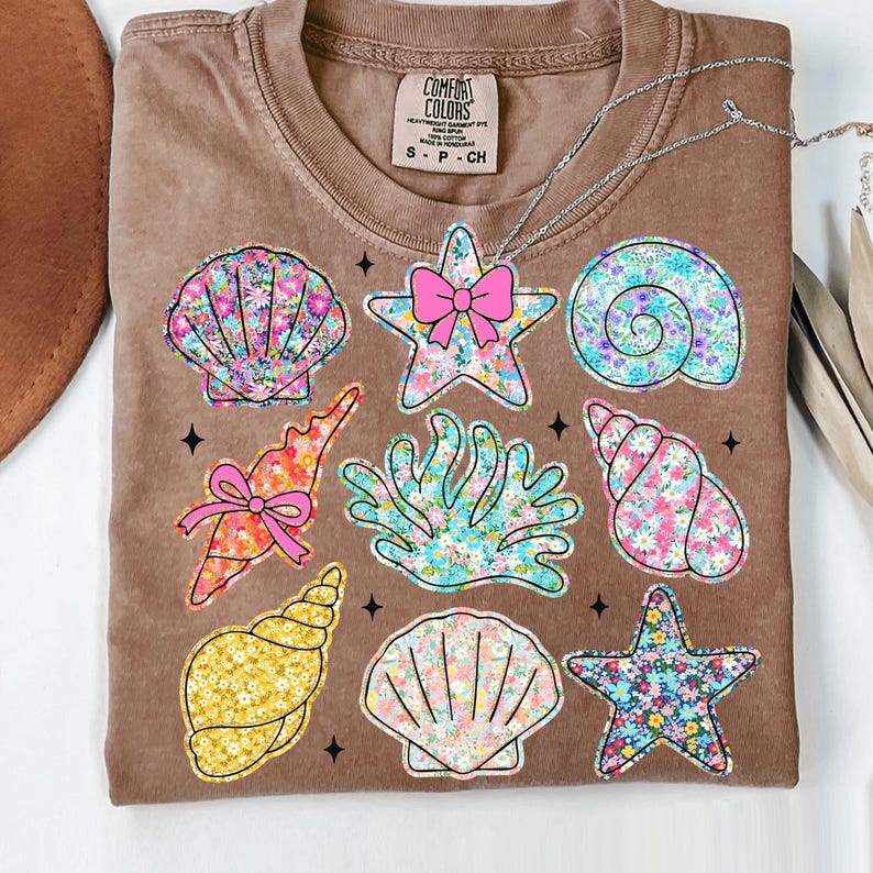 May include: A brown t-shirt with a floral pattern design featuring seashells, starfish, and coral. The text "COMFORT COLORS" is printed on the shirt.