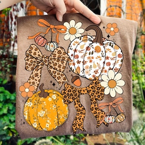May include: A brown t-shirt featuring a fall-themed design. The design includes two pumpkins, one orange and one white, with floral patterns. There are also leopard print bows, daisies, and small hanging gourds. The overall aesthetic is autumnal.