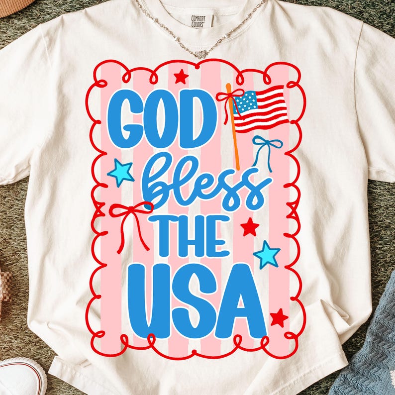Puede incluir: Camiseta blanca con un dise&ntilde;o patri&oacute;tico. El dise&ntilde;o presenta el texto "GOD bless THE USA" en azul, con un fondo a rayas rojas y rosas, estrellas y una bandera estadounidense.