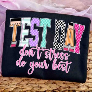 May include: Black t-shirt featuring the words "TEST DAY" in colourful block letters, and the phrase "don't stress do your best" in pink script. The letters are decorated with pencil and notebook paper designs.