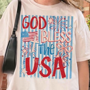 May include: White t-shirt with a patriotic design featuring the text "GOD BLESS The USA" in red and blue. The design includes an American flag, fireworks, and stars on a blue and white striped background.
