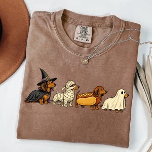 May include: A brown t-shirt featuring a graphic of four dachshunds in Halloween costumes: a witch, a mummy, a hot dog, and a ghost. The shirt has a crew neckline and a Comfort Colors brand label printed on the inside neck area with size S.