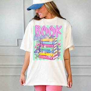 Puede incluir: Una camiseta de manga corta color hueso con un estampado gr&aacute;fico de una pila de libros coloridos y el texto "BOOK lover" en rosa y azul. El dise&ntilde;o incluye ilustraciones de margaritas blancas y flores moradas alrededor de los libros.