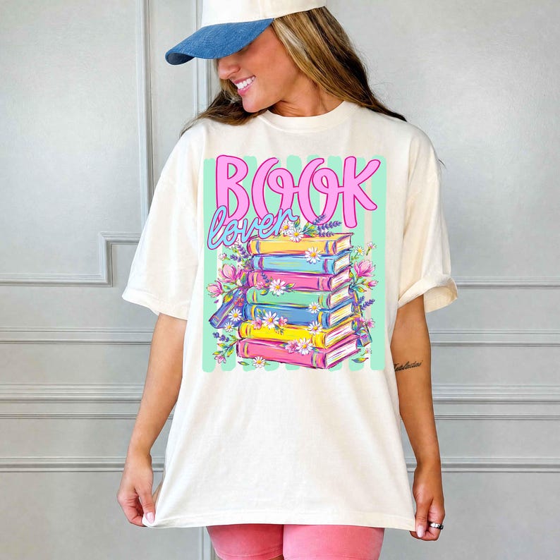 Puede incluir: Una camiseta de manga corta color hueso con un estampado gr&aacute;fico de una pila de libros coloridos y el texto "BOOK lover" en rosa y azul. El dise&ntilde;o incluye ilustraciones de margaritas blancas y flores moradas alrededor de los libros.