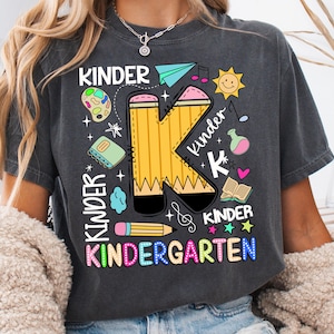 Pu&ograve; includere: T-shirt grigio scuro con un design colorato "Kindergarten". Il design presenta una grande "K" gialla a forma di matita con illustrazioni a tema scolastico, tra cui un aeroplano di carta, note musicali e un sole.