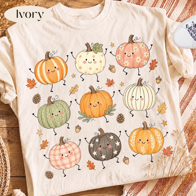 Puede incluir: Una camiseta de color marfil con un estampado gr&aacute;fico de nueve calabazas de dibujos animados con caras sonrientes, brazos y piernas. Las calabazas muestran varios patrones que incluyen rayas, lunares, flores y estrellas, intercalados con peque&ntilde;as bellotas, pi&ntilde;as y hojas.