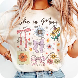 May include: A white t-shirt with the text "she is Mom" and various floral designs and bible verses. The verses include "Phil 4:13", "I Thess 1:9", "Rom 8:13", "Ecel 3:11", "Mark 10:27", "Jer 31:3", and "Matt 28:20".