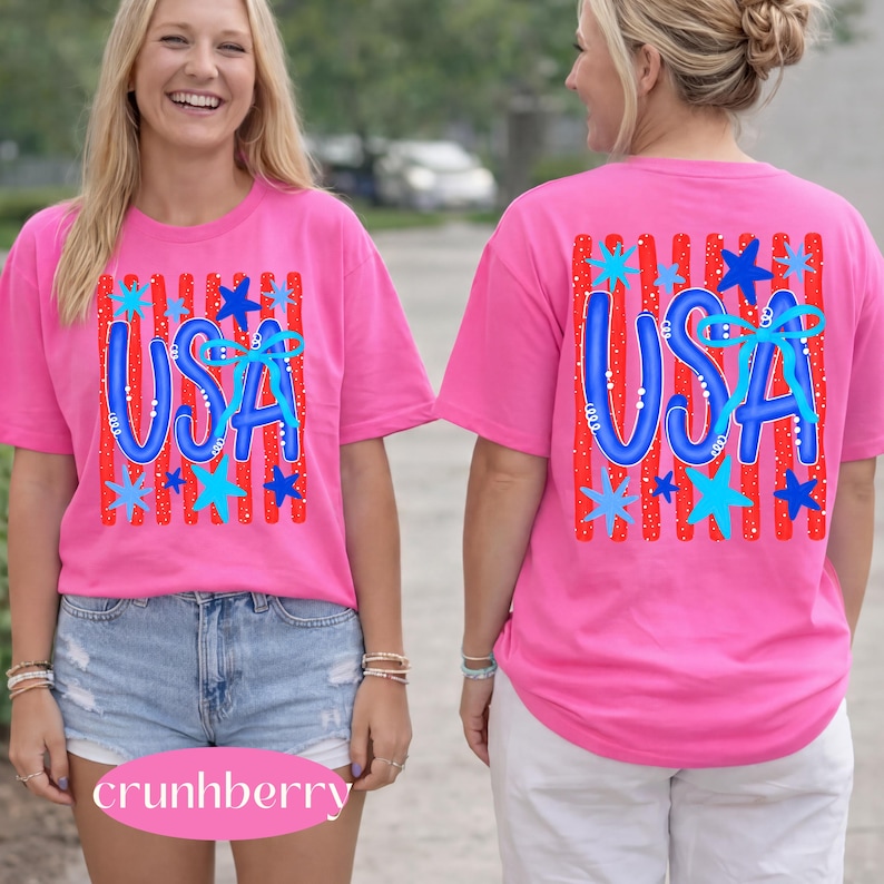 May include: A bright pink t-shirt with the word "USA" in blue, decorated with blue stars and red stripes. The shirt is a short-sleeved, casual style, perfect for patriotic events.