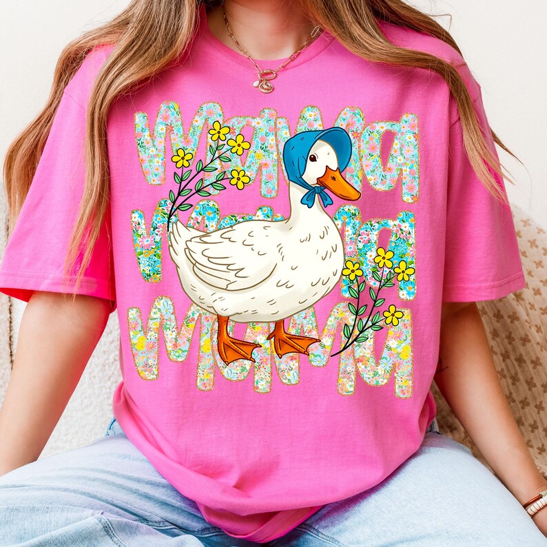 May include: A pink t-shirt with a playful design. The graphic shows the word "MAMA" in a floral pattern, framing a cartoon duck wearing a blue bonnet and a sprig of yellow flowers.