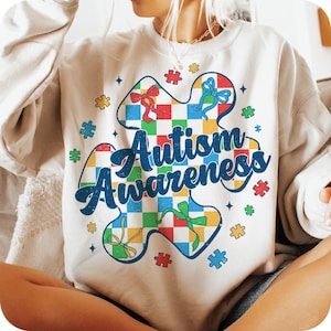 May include: A white sweatshirt with a colorful puzzle piece design that says "Autism Awareness". The puzzle piece is made up of squares with different colors and has bows on each corner.