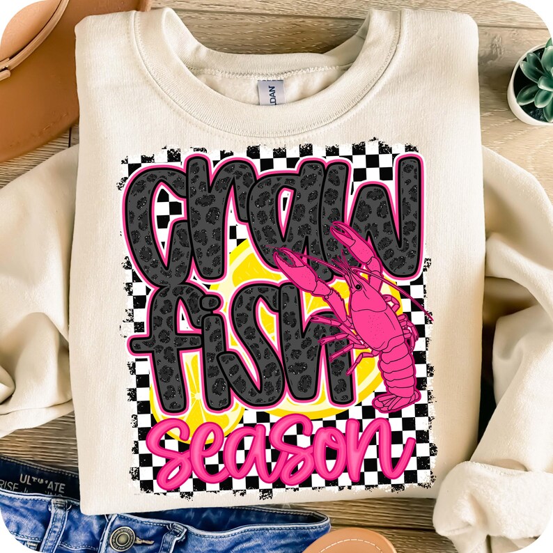 May include: A cream-colored sweatshirt with a black and white checkered background. The text "Crawfish Season" is written in black with a leopard print pattern. A pink crawfish is on the right side of the text.