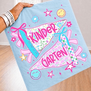 Peut inclure: Un tissu bleu clair avec une impression graphique repr&eacute;sentant deux fanions. Le fanion sup&eacute;rieur affiche le mot KINDER en rose, et le fanion inf&eacute;rieur affiche le mot GARTEN en rose. Le motif inclut des rubans roses, des &eacute;toiles, des fleurs et des smileys.